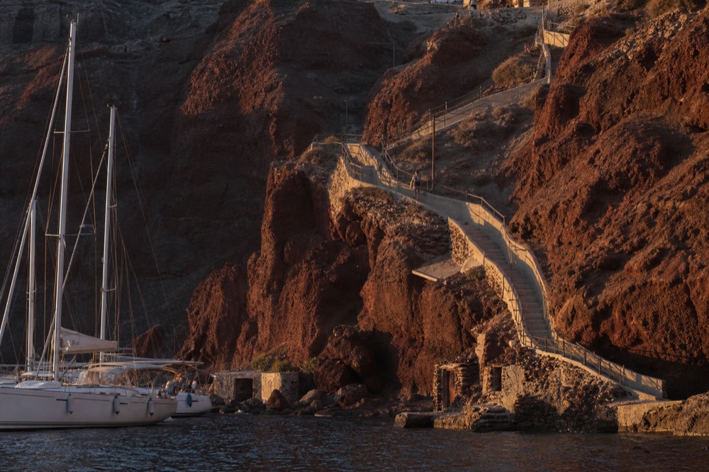 A stairway of worn switchback steps climbing a red cliff face from a tiny harbour, iron handrail throwing a long shadow in late light
