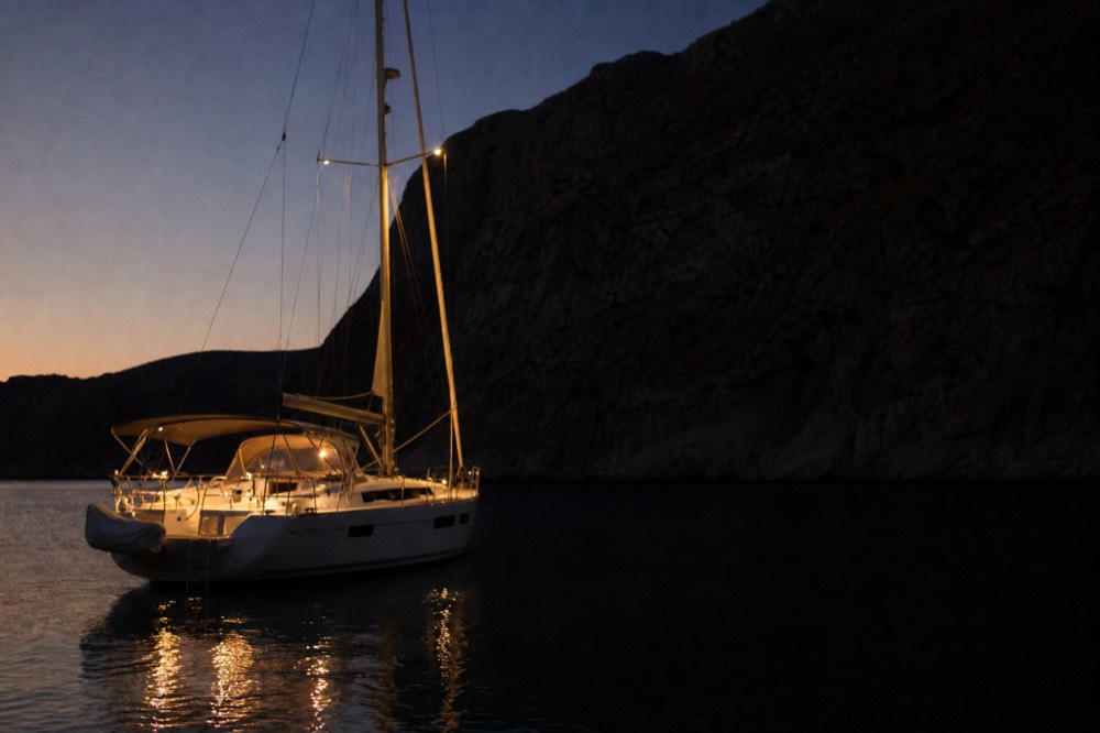 A yacht at anchor with deck lights burning during blue hour, mirrored on flat black water beneath a dark cliff silhouette