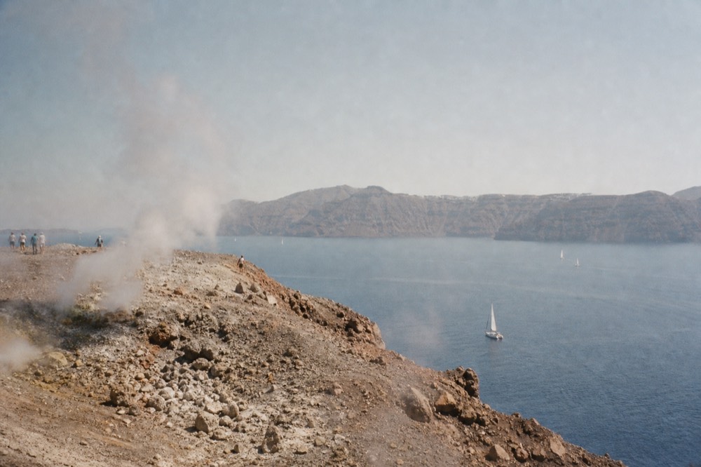 A steaming rocky crater rim of grey and ochre volcanic rubble under hard midday light, with the flat blue caldera and distant cliffs beyond