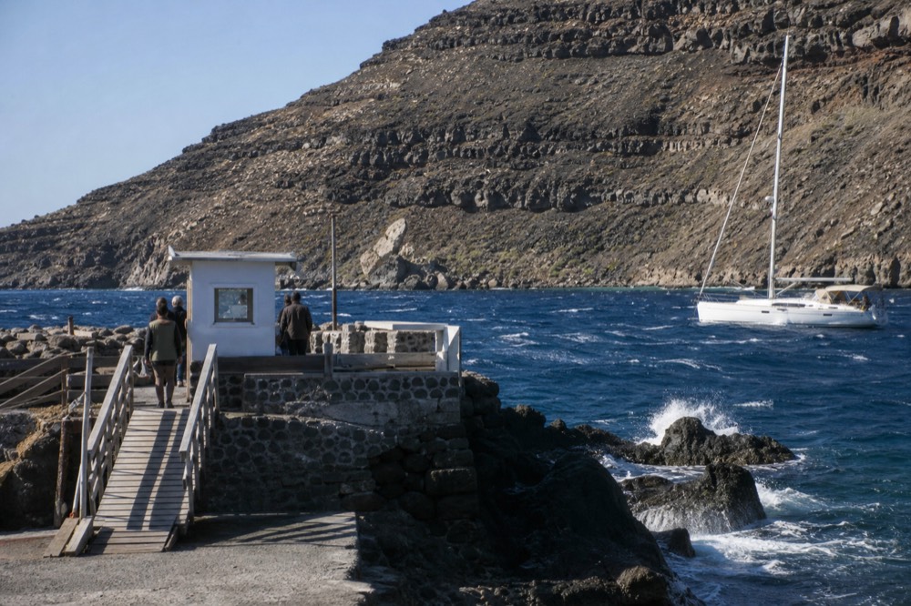 A rough stone landing jetty on black lava rock with a wooden gangway and a small ticket hut, bare volcanic slope rising steeply behind
