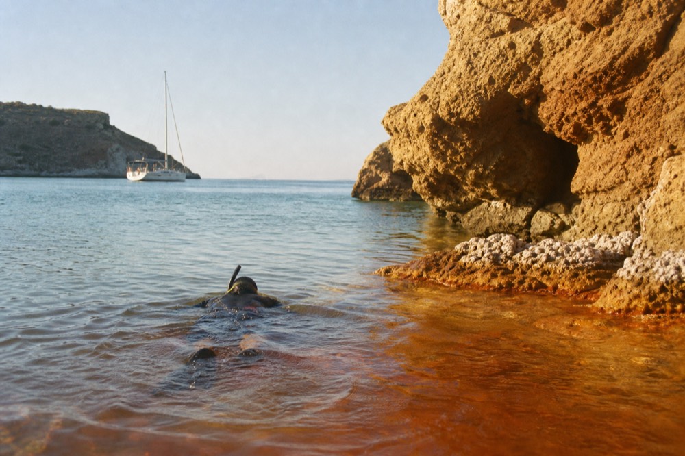 A snorkeller in a dark suit swimming through rust-coloured mineral water toward warm shallows under low ochre rock, clear blue sea behind